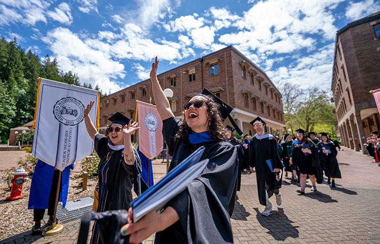 Graduates walking across campus holding flags, wearing caps and gowns and waving their hands in the air